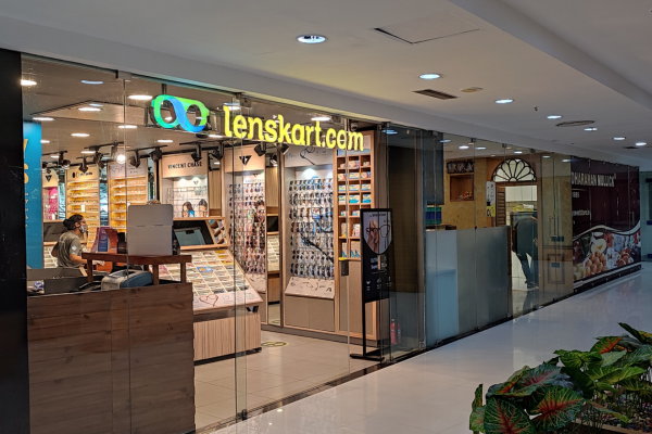 The image shows a LensKart store inside a mall or shopping complex. A person wearing a face mask is visible at a counter on the left. The store has large glass windows and is brightly lit, showcasing various eyeglasses and sunglasses on display racks. The brand's logo is prominently featured above the entrance.