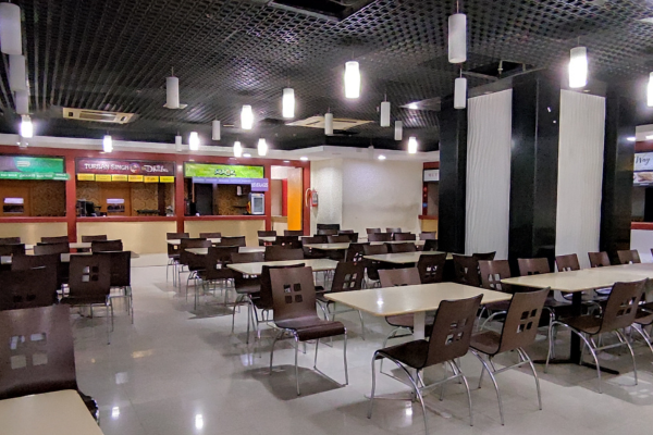 The image shows the interior of a large food court, which is a type of retail space dedicated to food and dining. The area is filled with rows of empty tables and chairs, with a variety of food stalls visible along the back wall, each with its own sign. The ceiling has a dark, grid-like pattern with a series of modern hanging light fixtures, and the floor is a light, reflective tile.