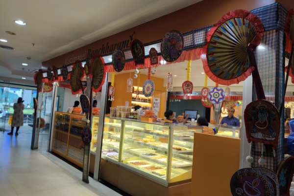 A well-lit Banchharam's sweet shop storefront at Infinity Group Retail mall, decorated with colorful traditional crafts.