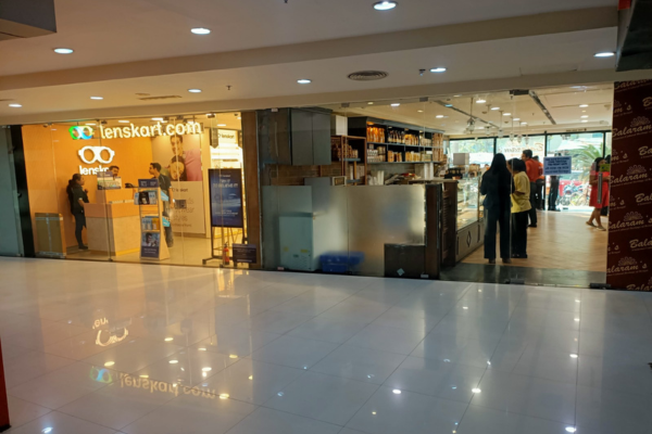 The image shows a LensKart store inside a mall or shopping complex. A person wearing a face mask is visible at a counter on the left. The store has large glass windows and is brightly lit, showcasing various eyeglasses and sunglasses on display racks. The brand's logo is prominently featured above the entrance.